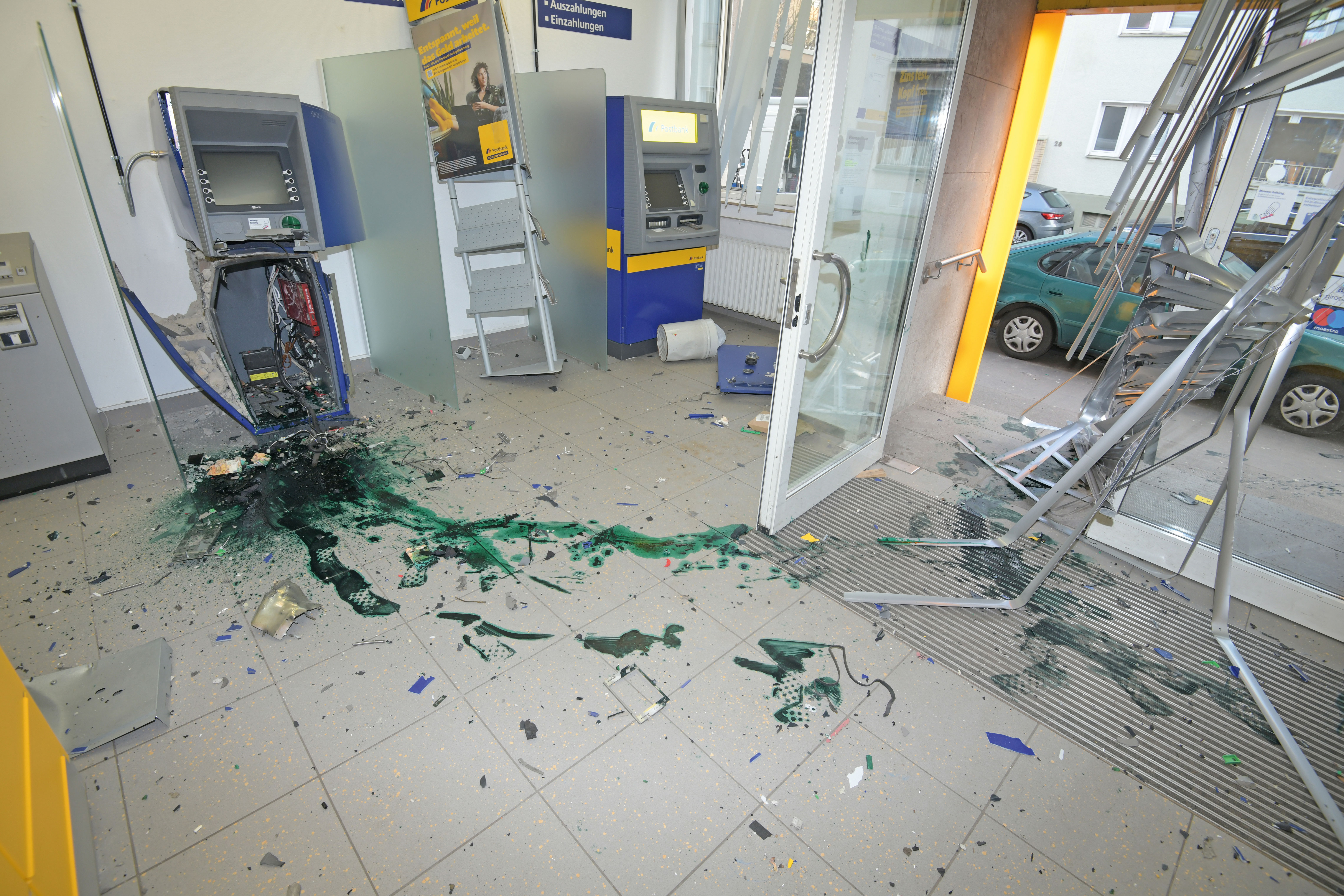 You can see a bank branch where an ATM has been blown up. There are many shattered pieces of the machine on the floor. There is a green ink stain in front of the machine and shoe prints made of green ink can be seen.