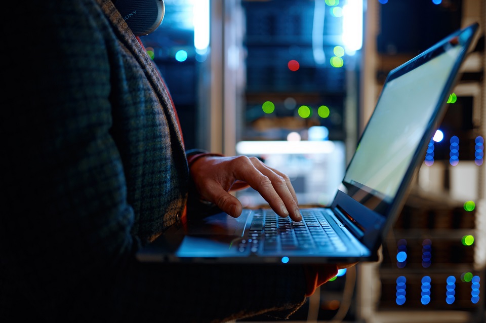 Ein Mann steht mit Laptop in einem Serverraum. Schemenhafte Darstellung
