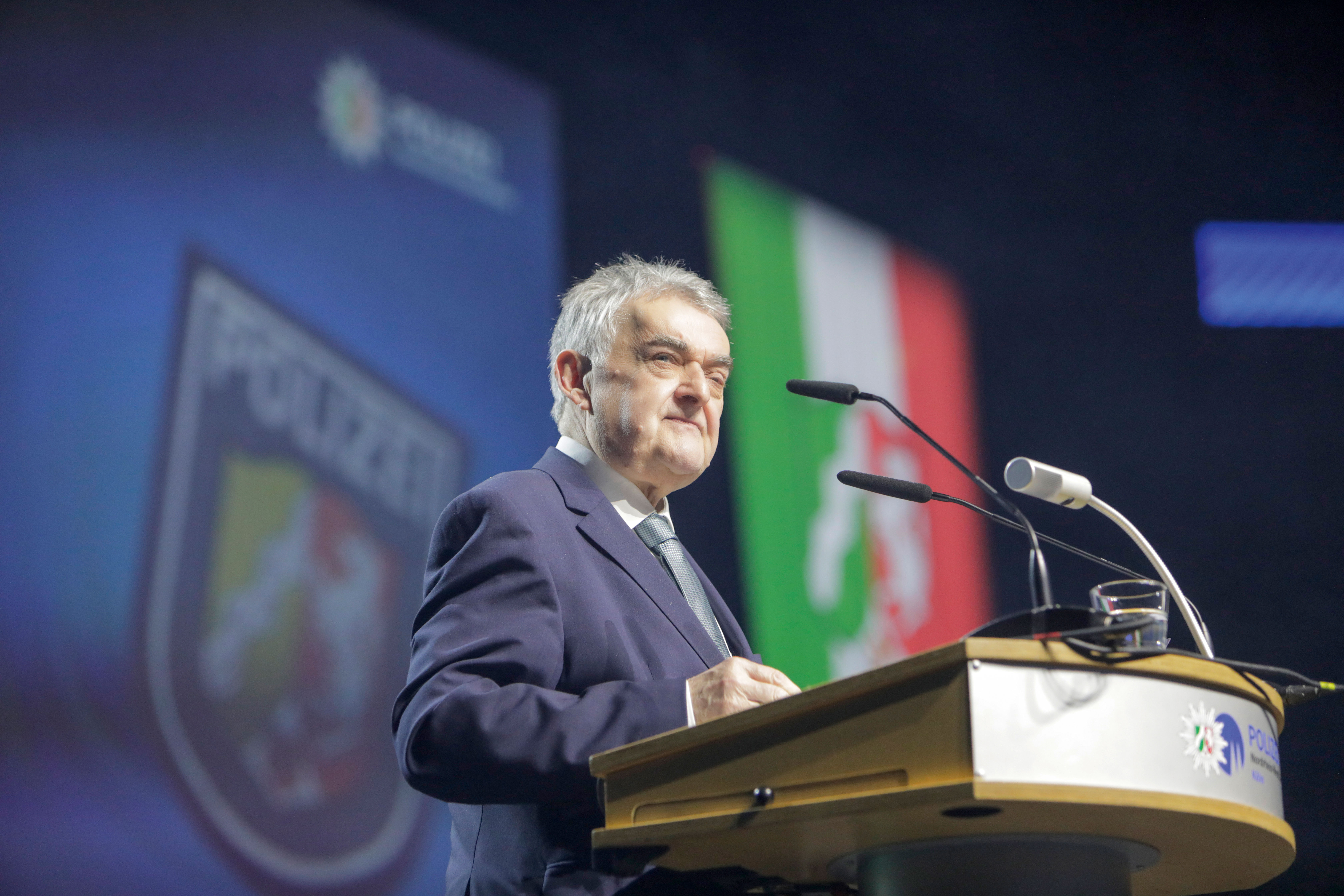 Innenminister Herbert Reul spricht an einem Podium. Im Hintergrund ist ein Banner der Polizei der Polizei NRW und die Flagge von NRW verschwommen zu erkennen.