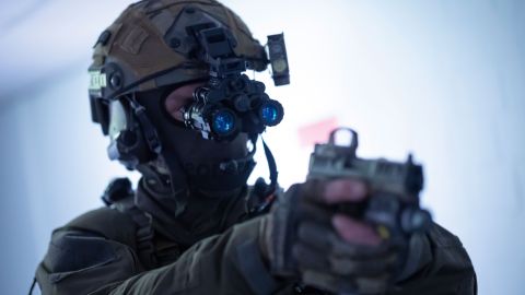 A special forces officer can be seen in a close-up up to his chest. He is wearing a helmet with a sight attached to it. He is holding a gun and pointing it in front of him.