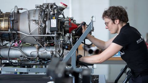 Prüferin Stefanie Bohr untersucht einen großen Motor, der aus vielen Teilen besteht. Im Hintergrund ist eine weiße Wand zu sehen.